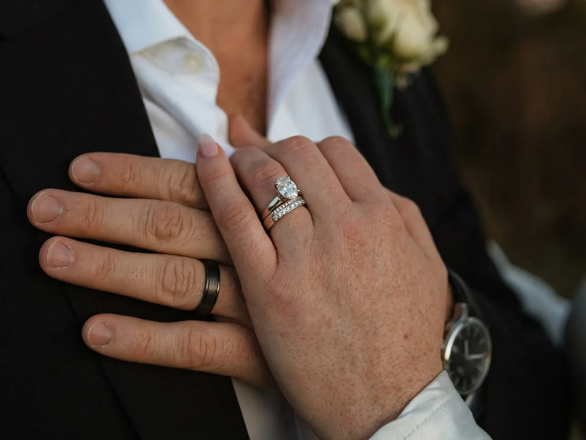 Hands with wedding rings, one with a diamond, resting on a suit.
