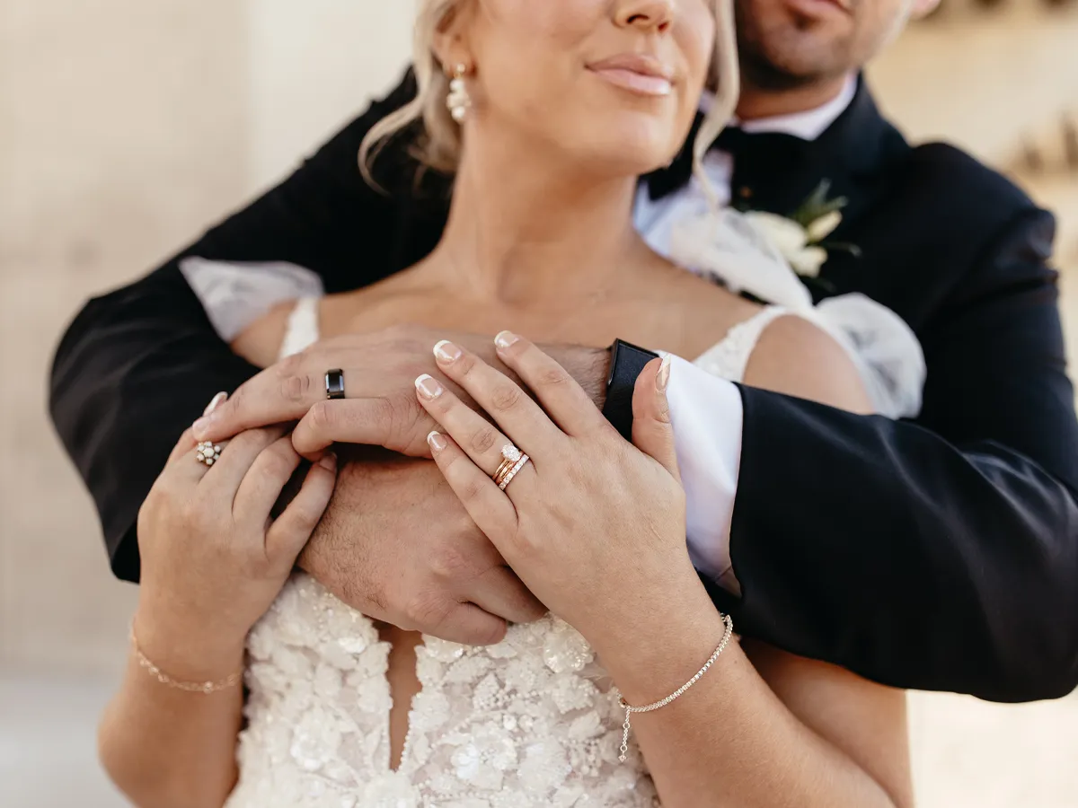 Bride and groom embrace, showcasing wedding rings and elegant attire.