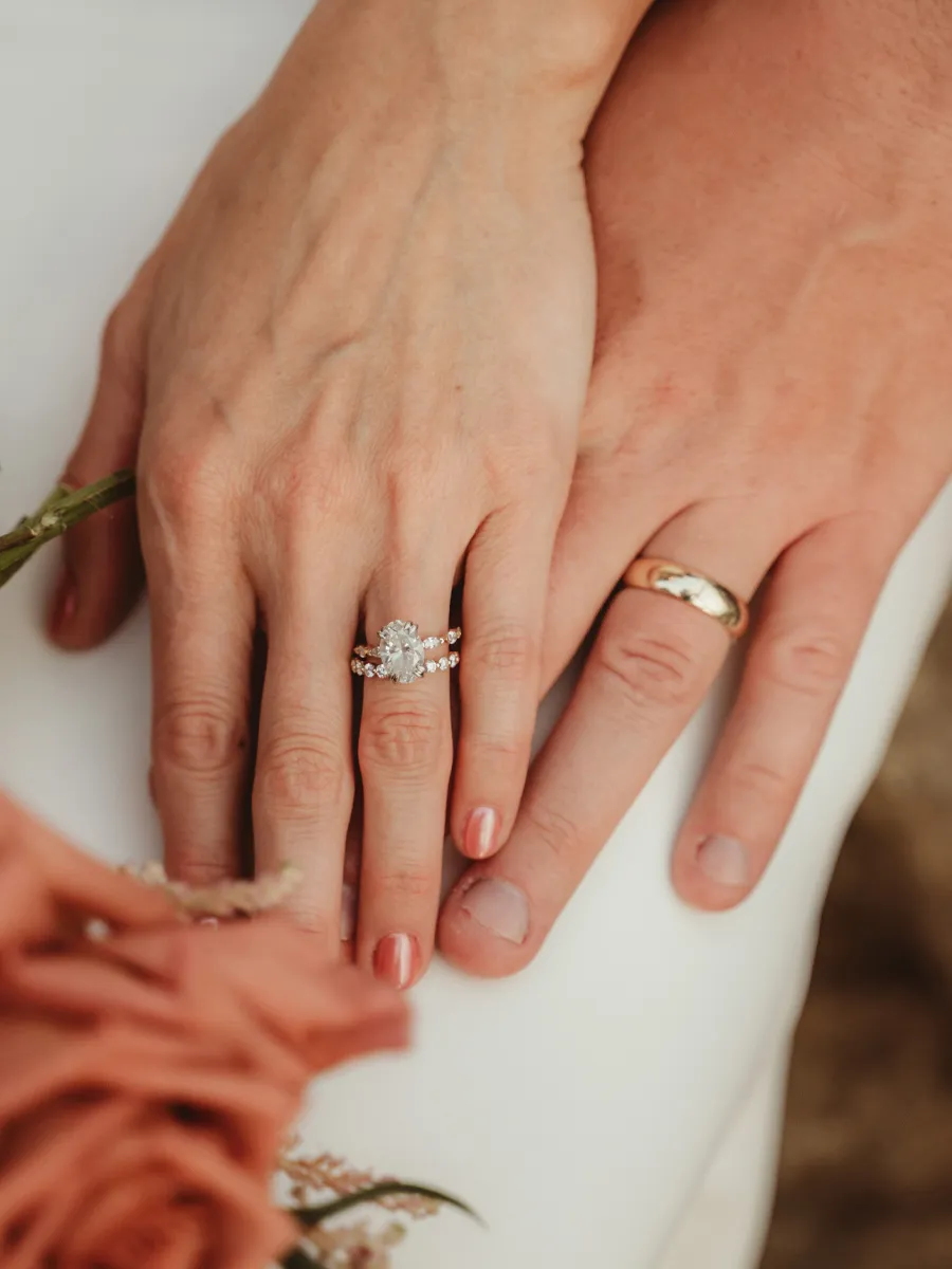 Close-up of two hands with wedding rings, one with a diamond engagement ring, over a white surface with flowers.