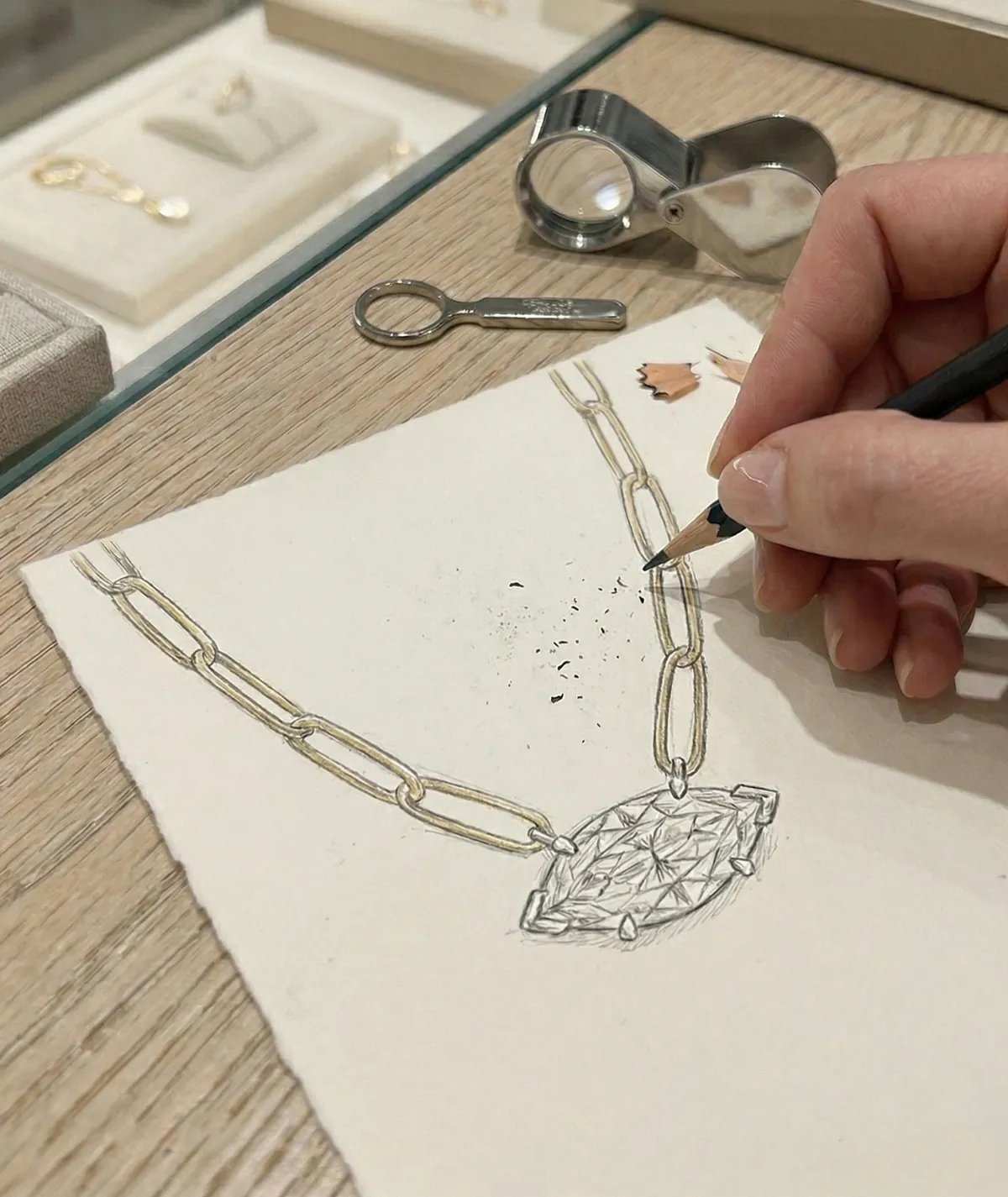 A hand sketches a necklace design with a pencil on paper, featuring a large gemstone pendant.
