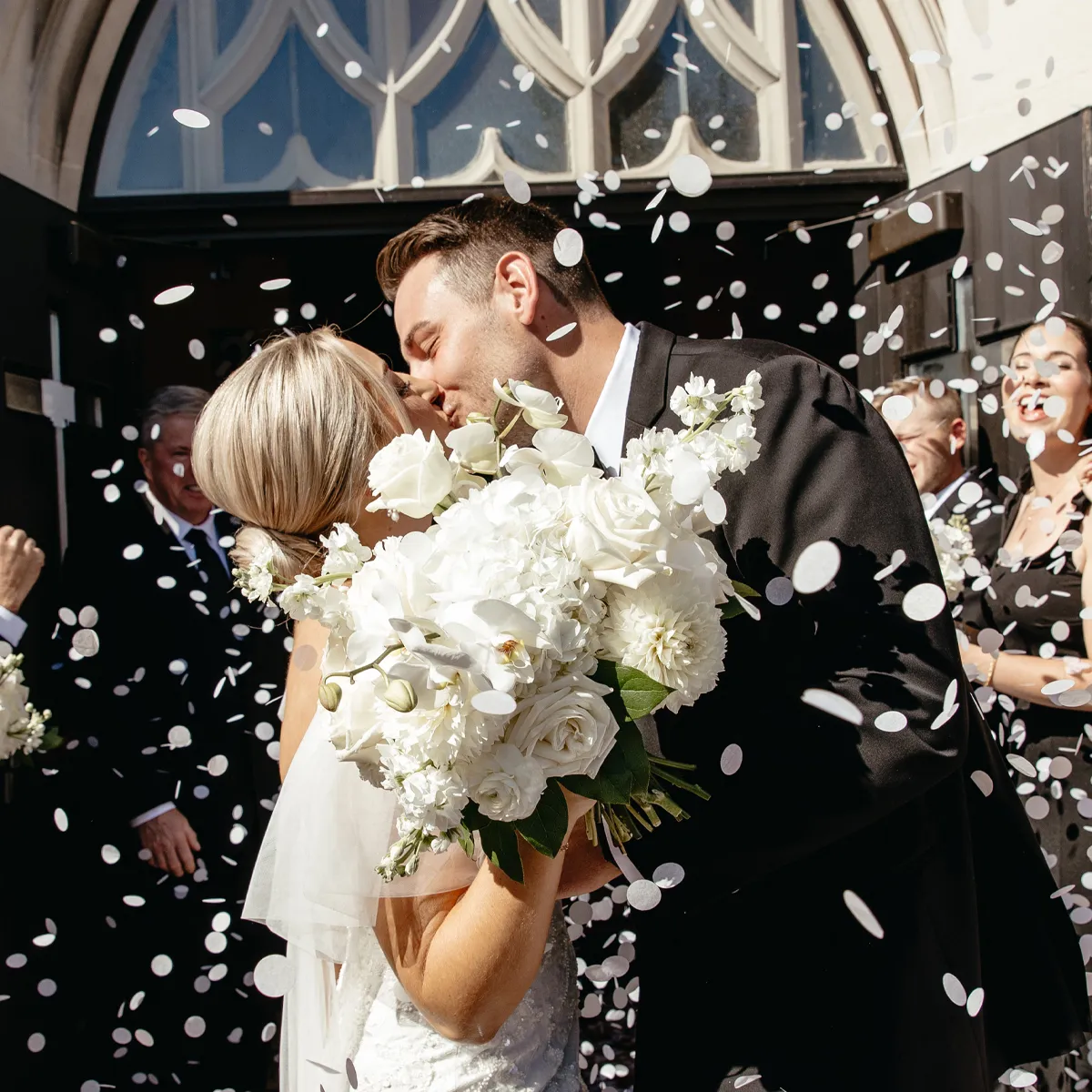 Bride and groom kiss under confetti outside a church, holding a bouquet of white flowers.