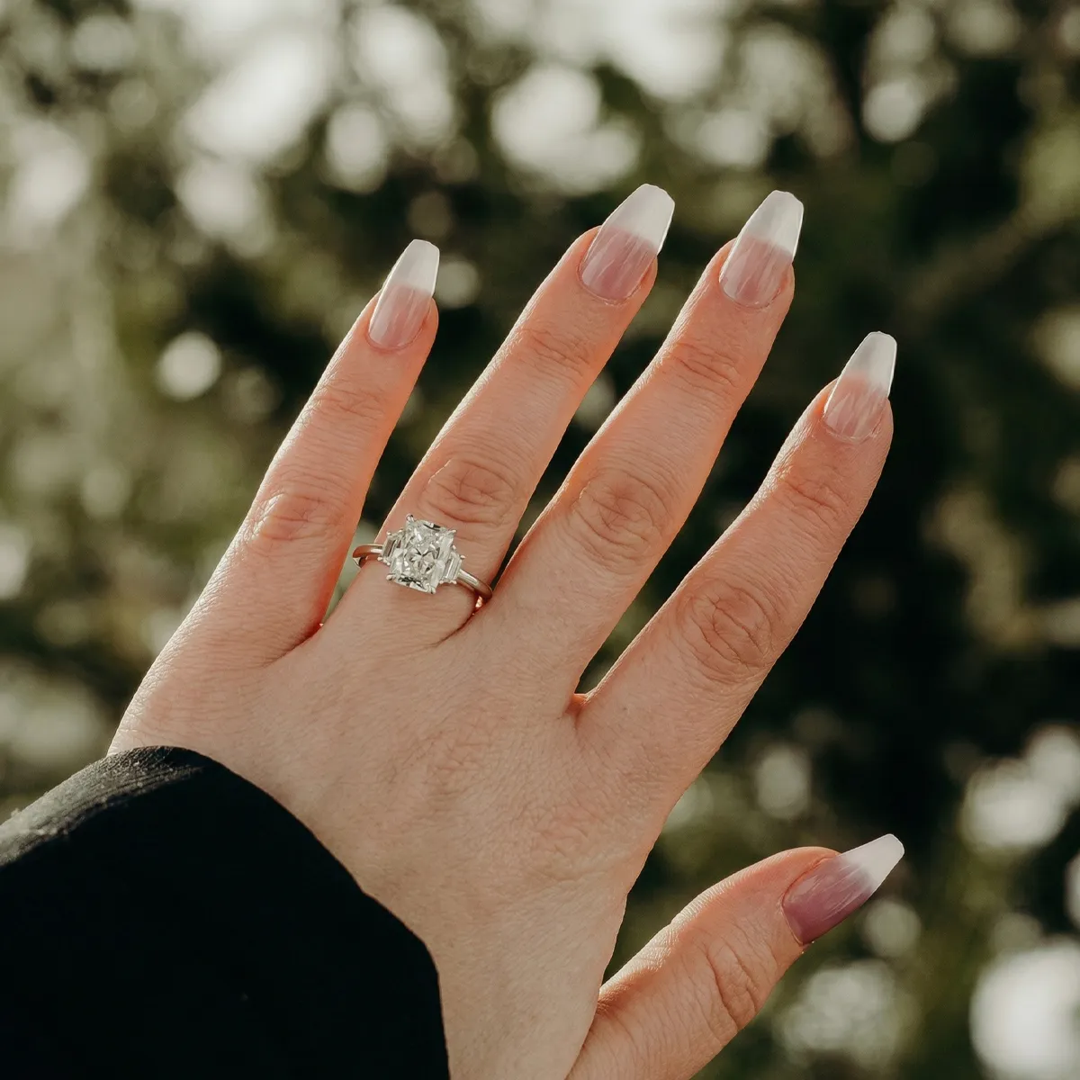 A hand with long nails displays a large diamond ring outdoors.