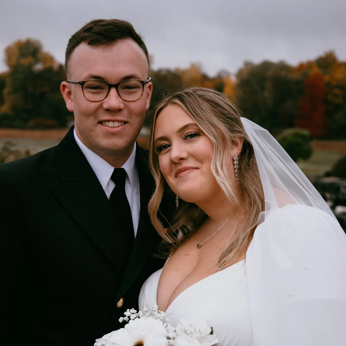 Bride and groom smiling outdoors with autumn trees in the background.