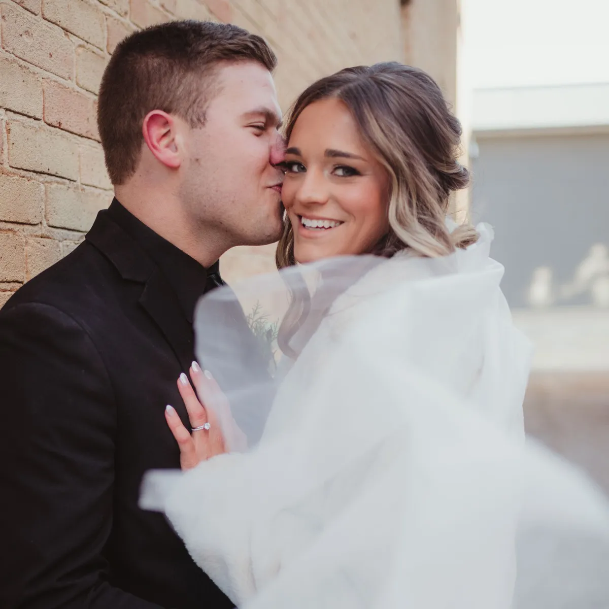 A couple embraces, with the man kissing the woman's cheek. She smiles, showing an engagement ring.