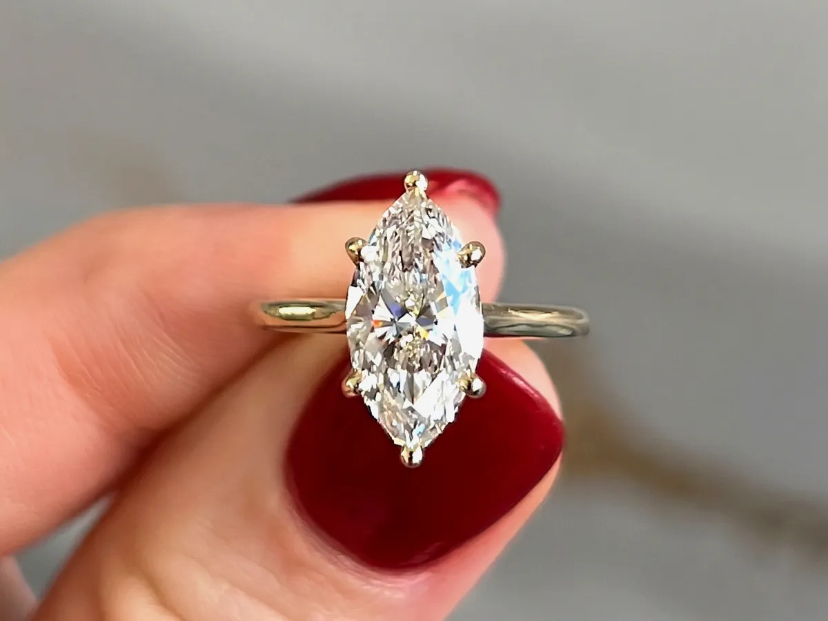 A hand holding a marquise-cut diamond ring with a gold band against a blurred background.
