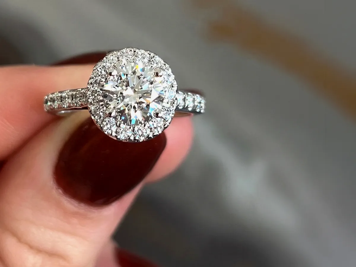 A hand holding a diamond engagement ring with a halo setting.