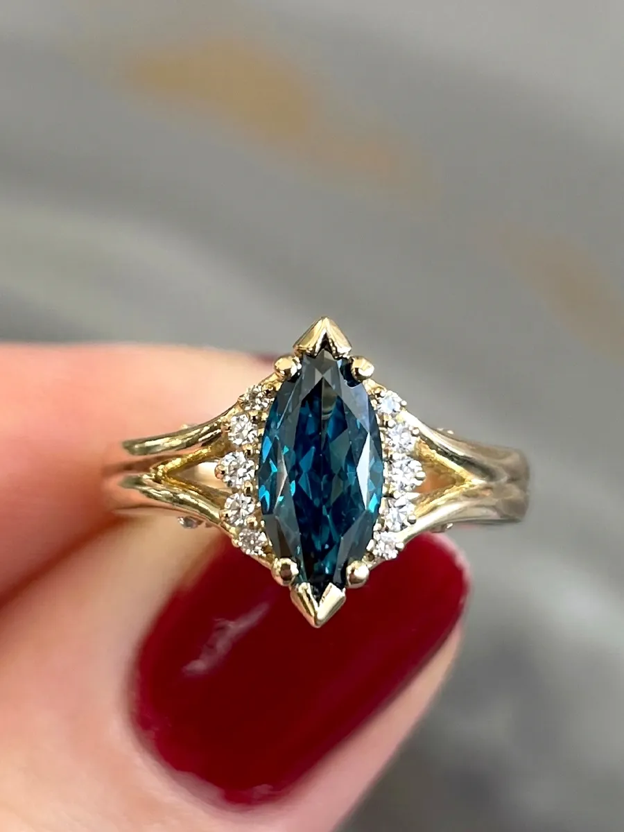 A marquise-cut blue gemstone ring with diamond accents held by a hand with red nail polish.