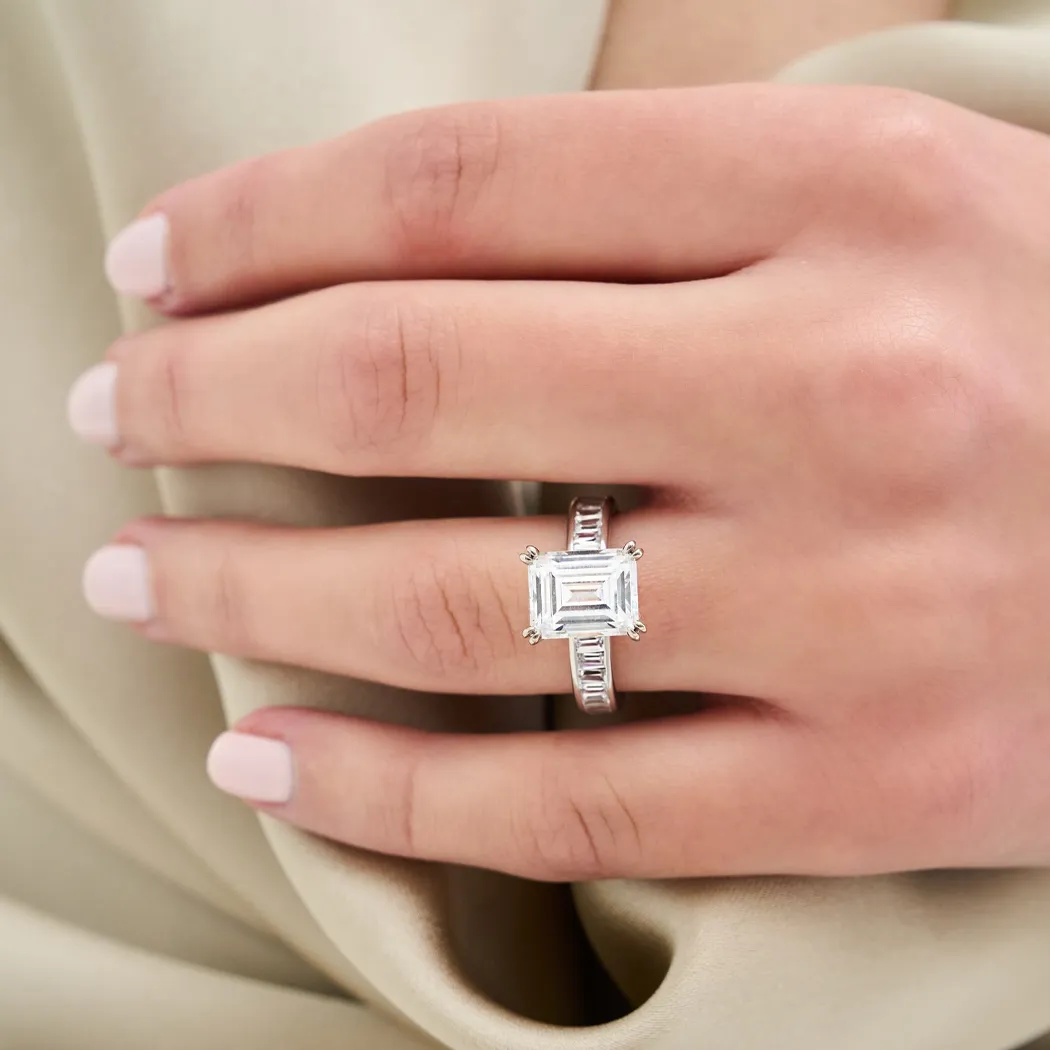 A hand with a square-cut diamond engagement ring on a light fabric background.