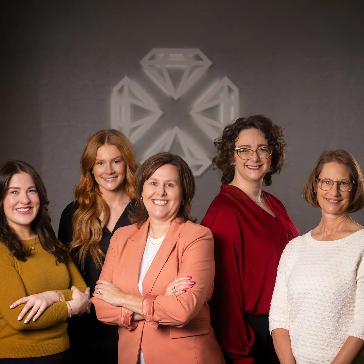 Five women smiling and posing together in front of a diamond logo on a wall.