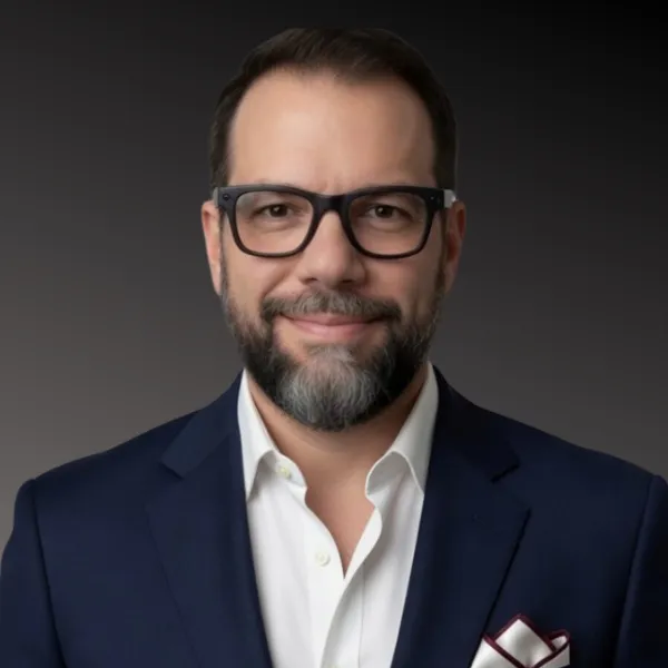 Man in blue jacket with white shirt, beard and glasses, smiling against a dark background.