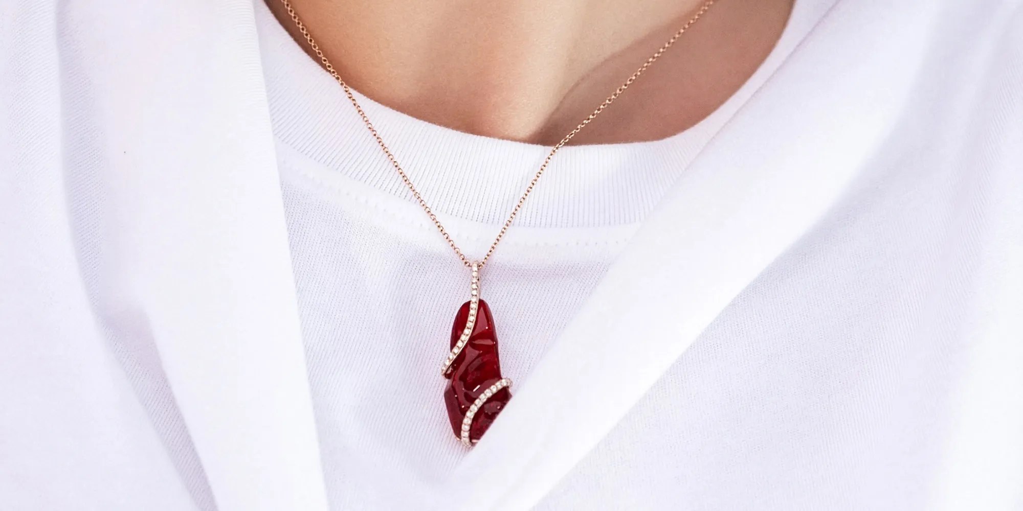 A red gemstone pendant with diamond accents on a gold chain, worn over a white shirt.