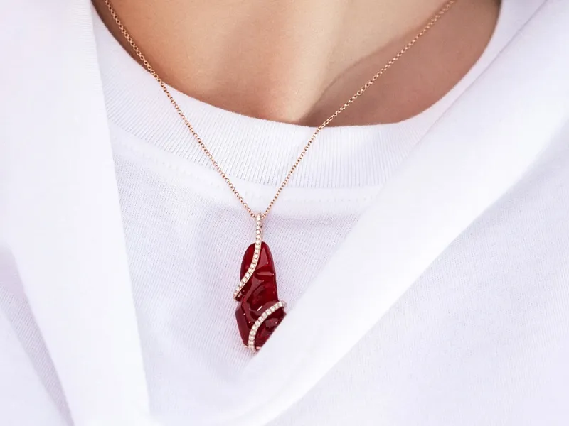 A red gemstone pendant with diamond accents on a gold chain, worn over a white shirt.