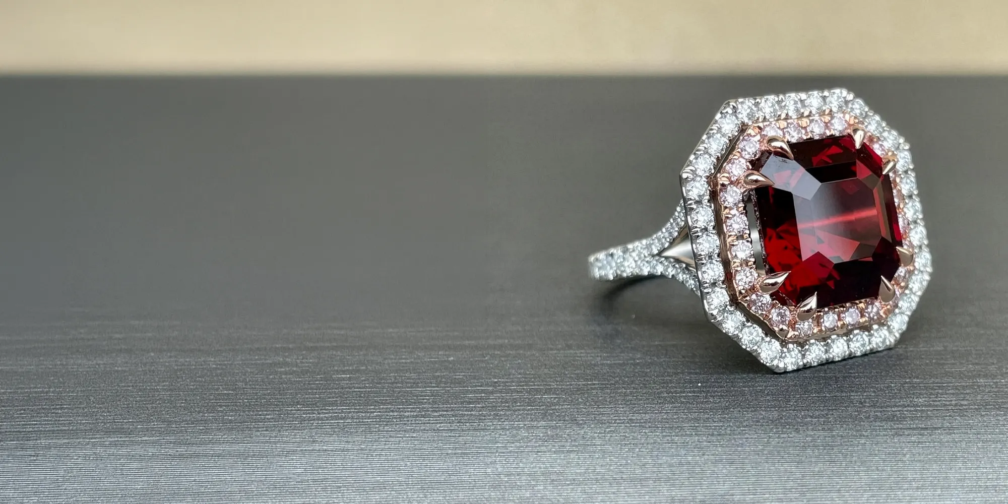 Red gemstone ring with diamond halo on a gray surface.