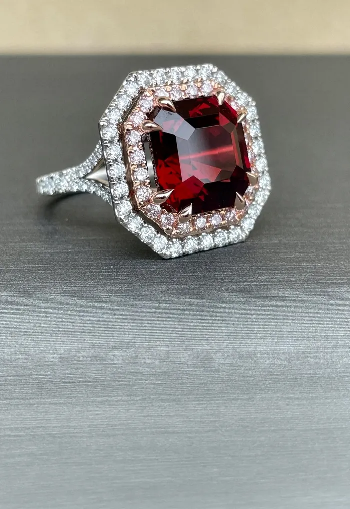 Red gemstone ring with diamond halo on a gray surface.