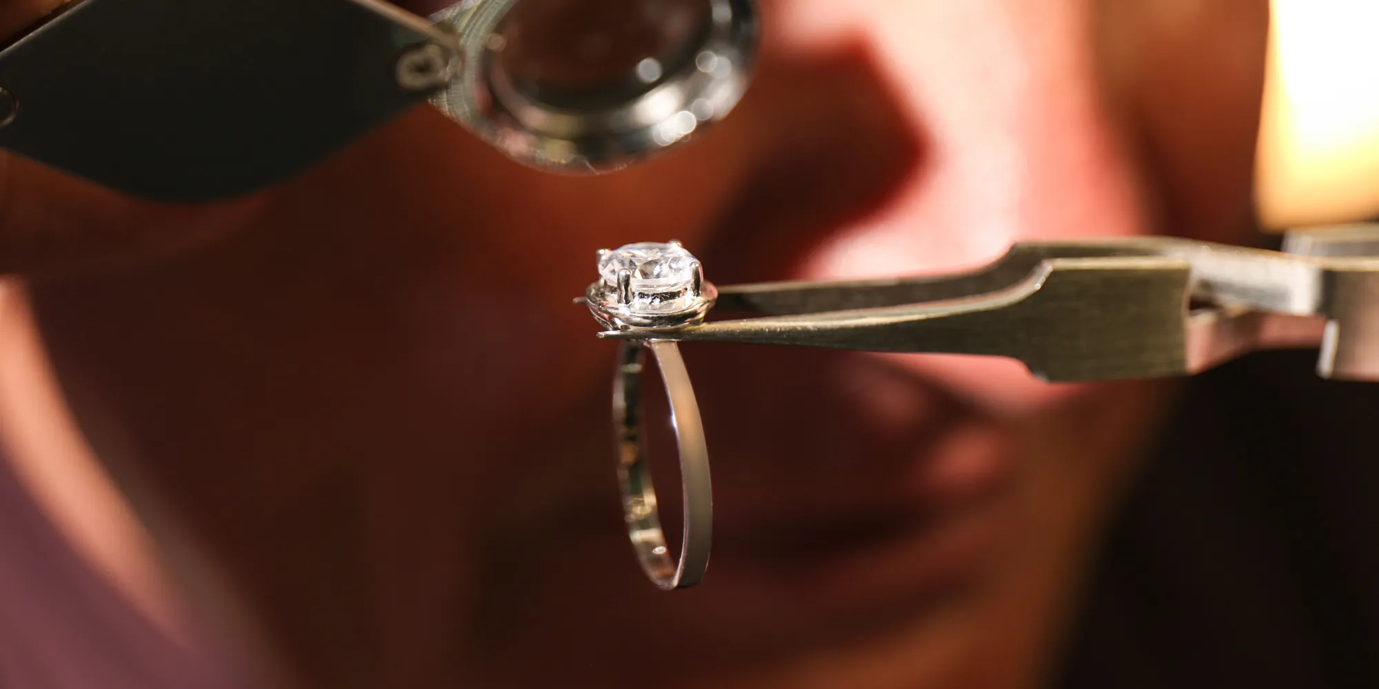 A jeweler inspects a diamond ring with a magnifying glass and tweezers.