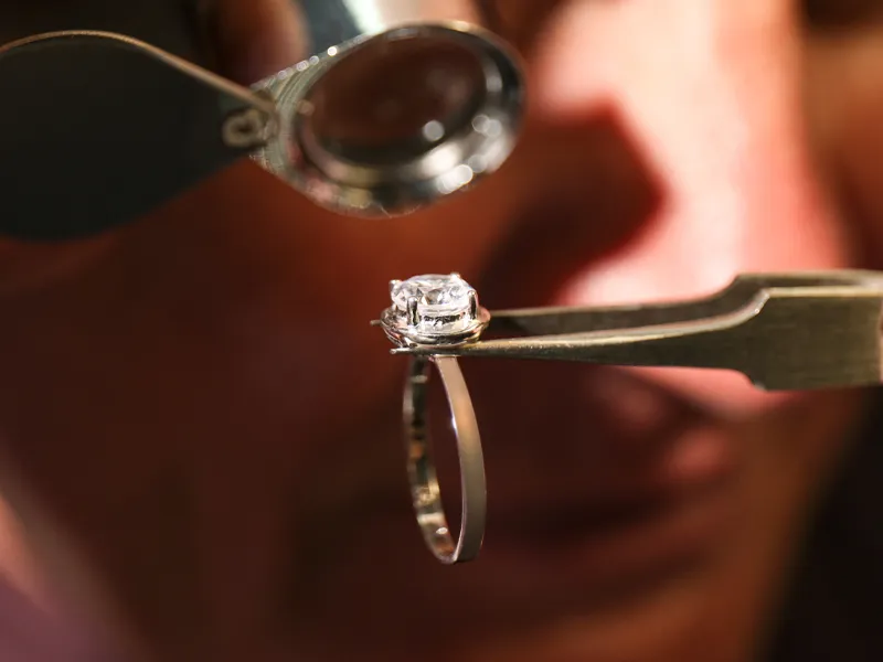 A jeweler inspects a diamond ring with a magnifying glass and tweezers.