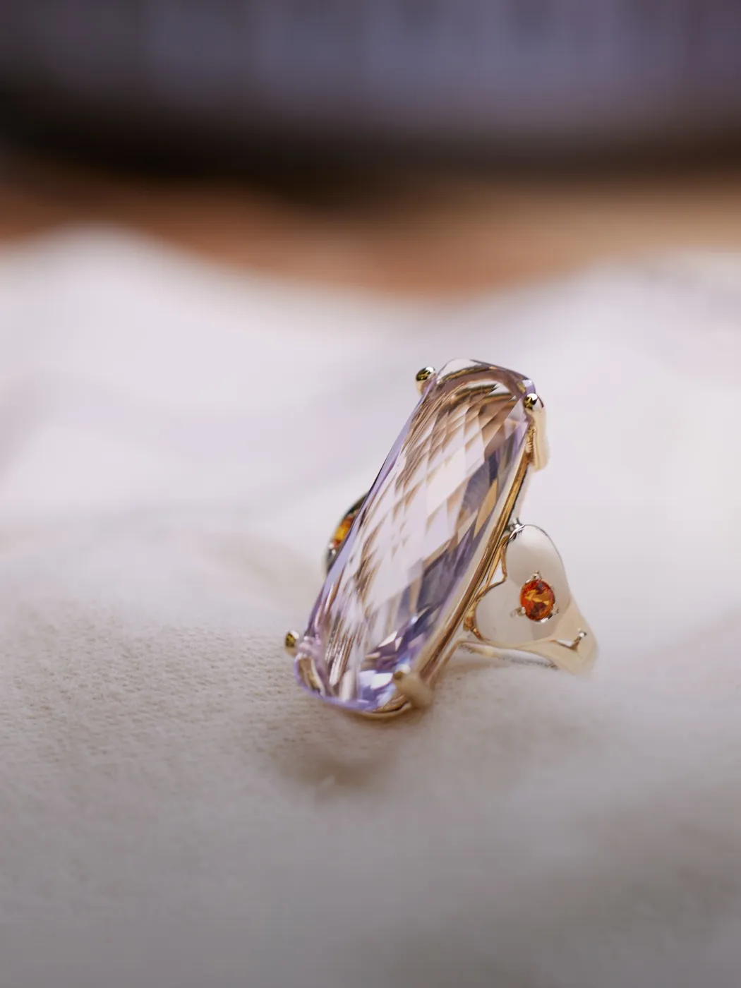 Elegant ring with a large, faceted gemstone and small orange accents on a soft fabric background.