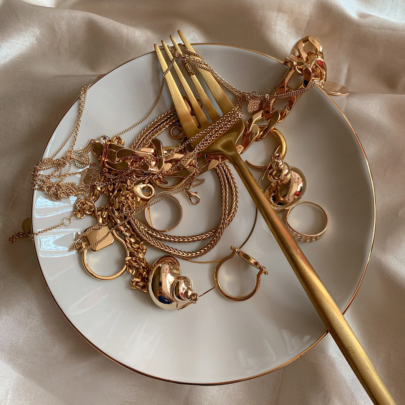A gold fork and assorted gold jewelry on a white plate with a satin background.