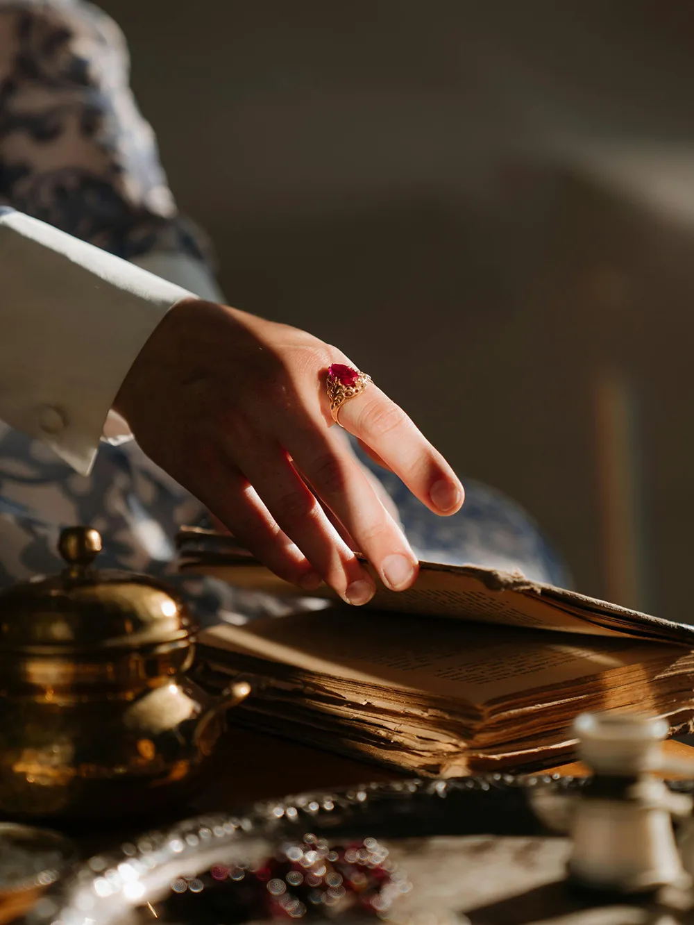 A hand with a red gemstone ring turns pages of an old book, with a brass teapot nearby.