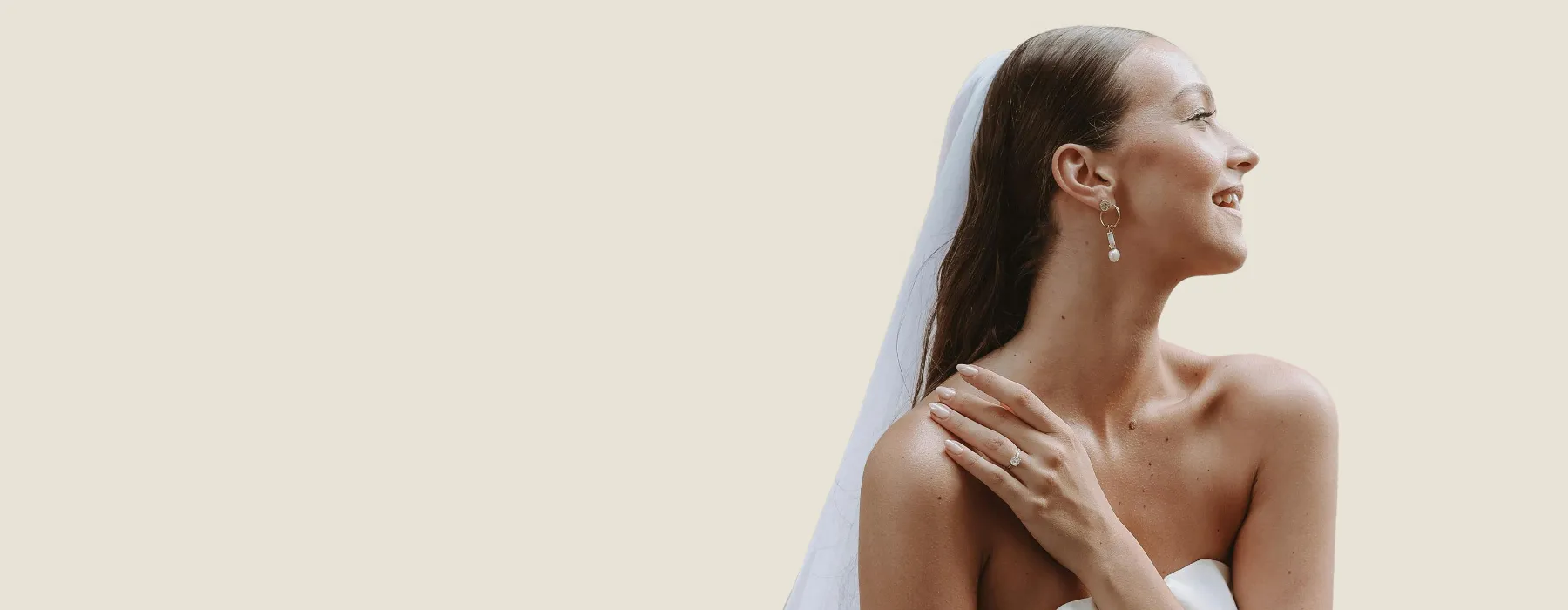 Bride smiling in profile, wearing a veil and earrings, with a ring on her hand against a light background.