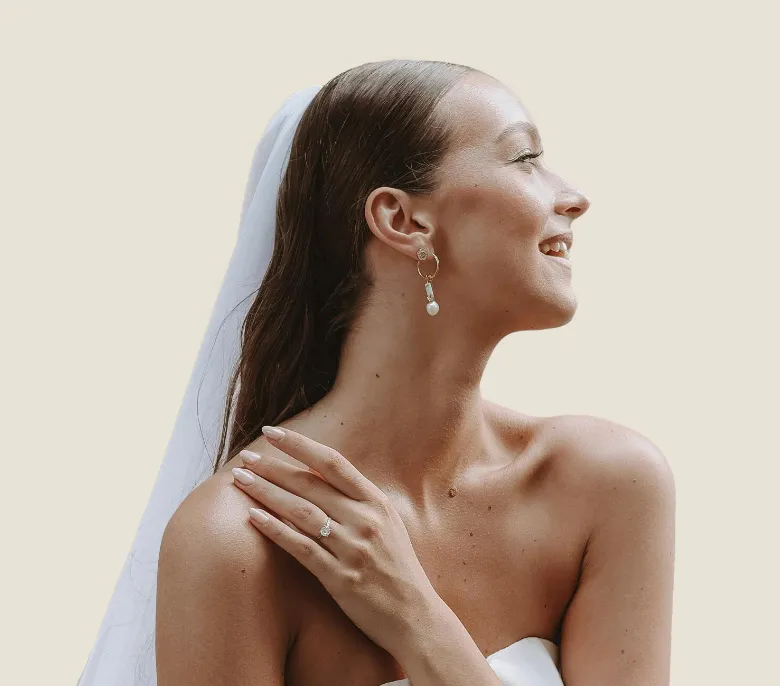 Bride smiling in profile, wearing a veil and earrings, with a ring on her hand against a light background.