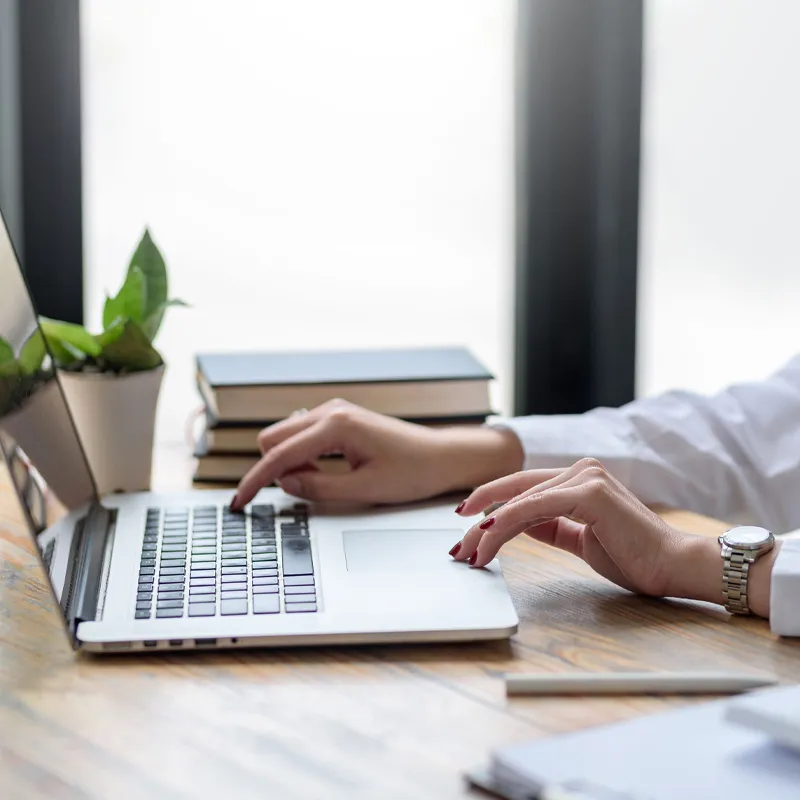 Person typing on a laptop with a plant and books in the background, representing a workspace.