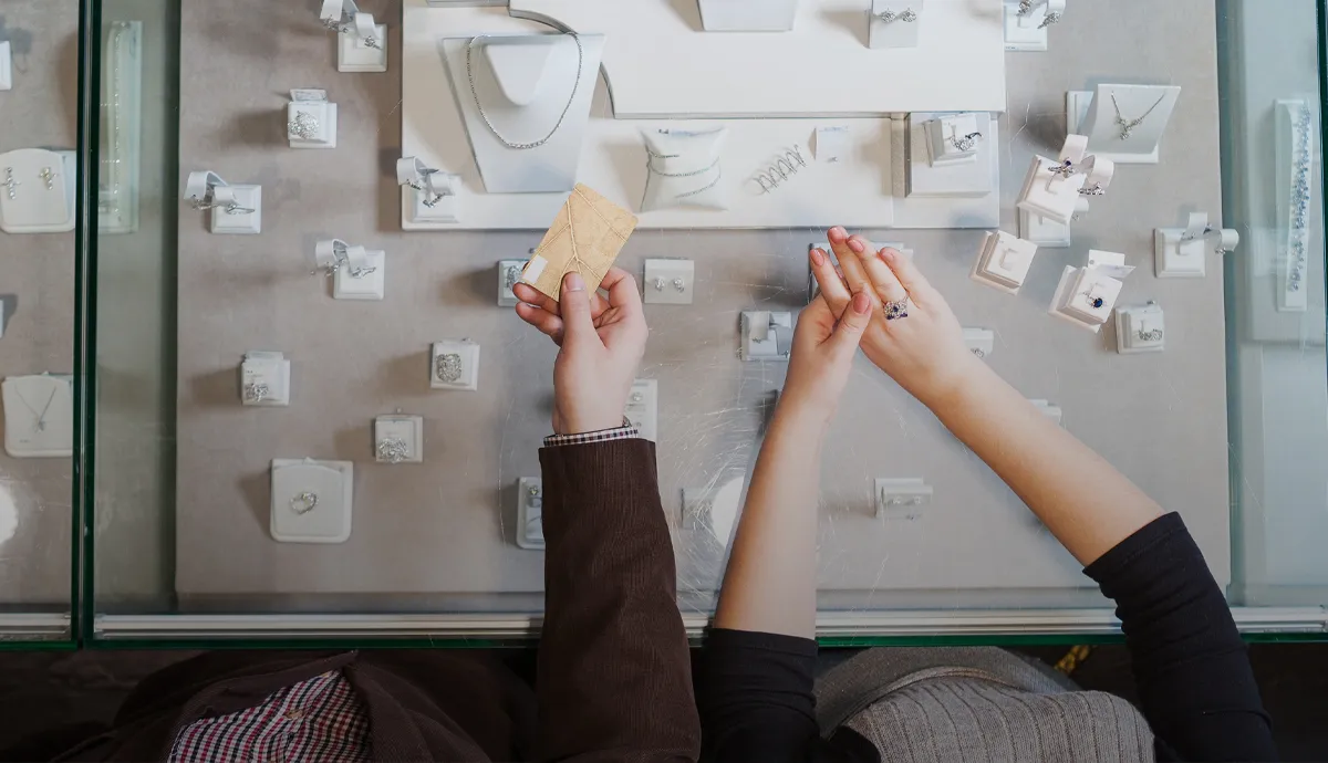 Two people examining jewelry in a store, one holding a card, the other showcasing a ring.