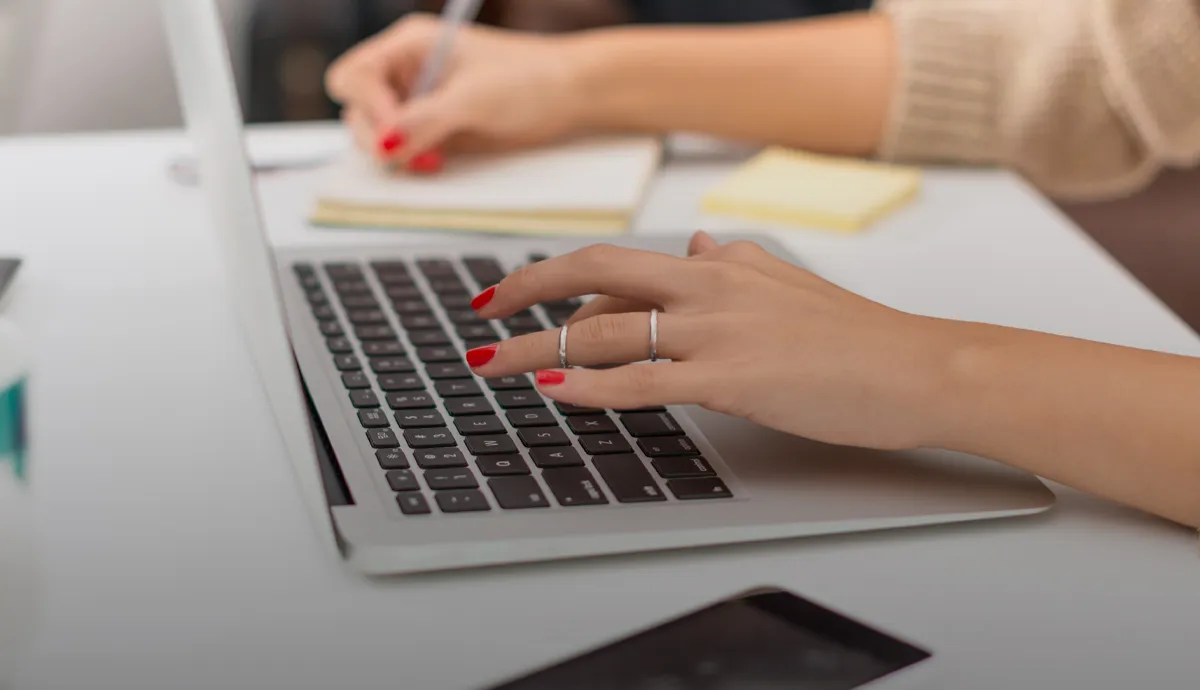 A hand with red nails typing on a laptop, while another hand writes notes on a notepad.