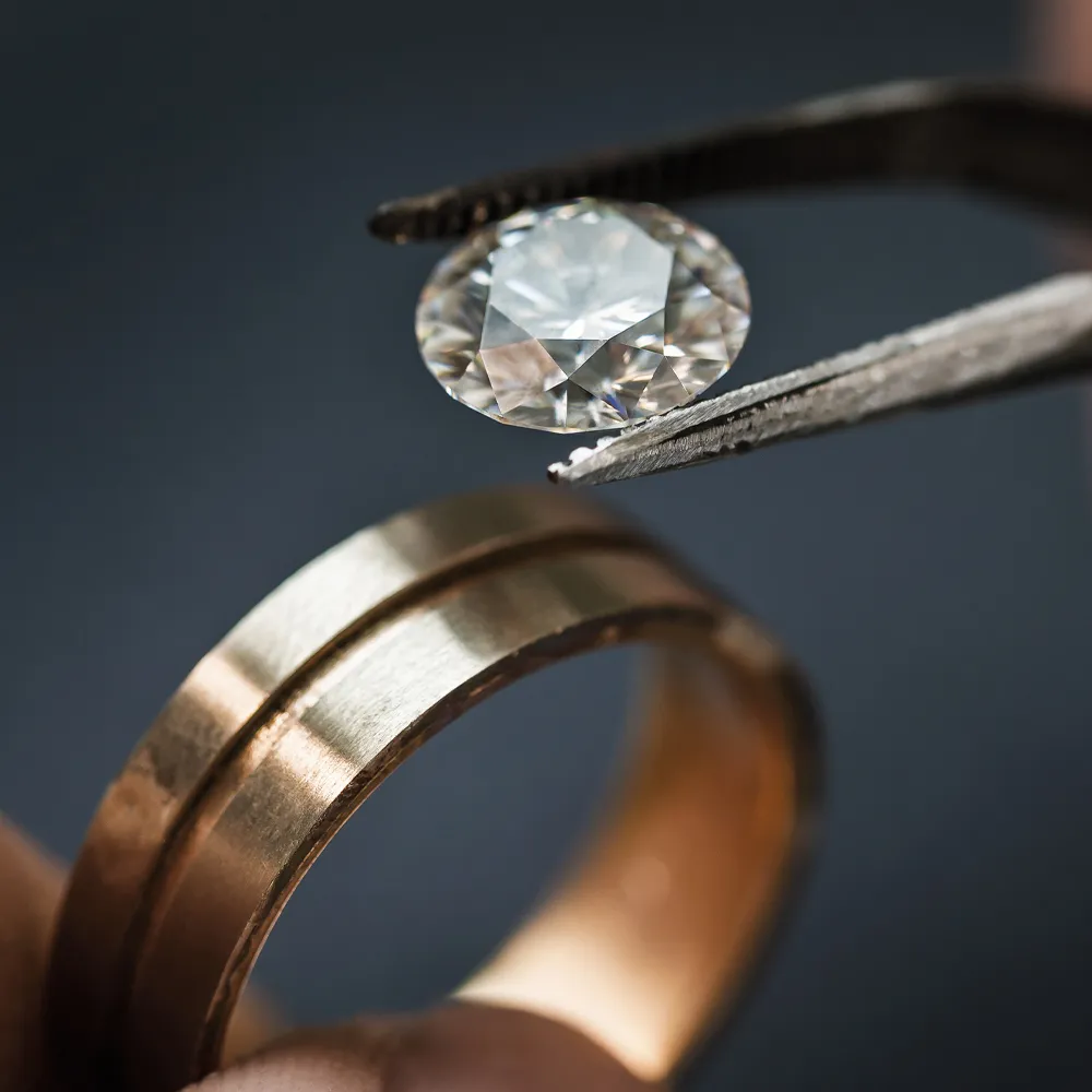 A diamond being held with tweezers above a gold wedding band, showcasing fine jewelry craftsmanship.