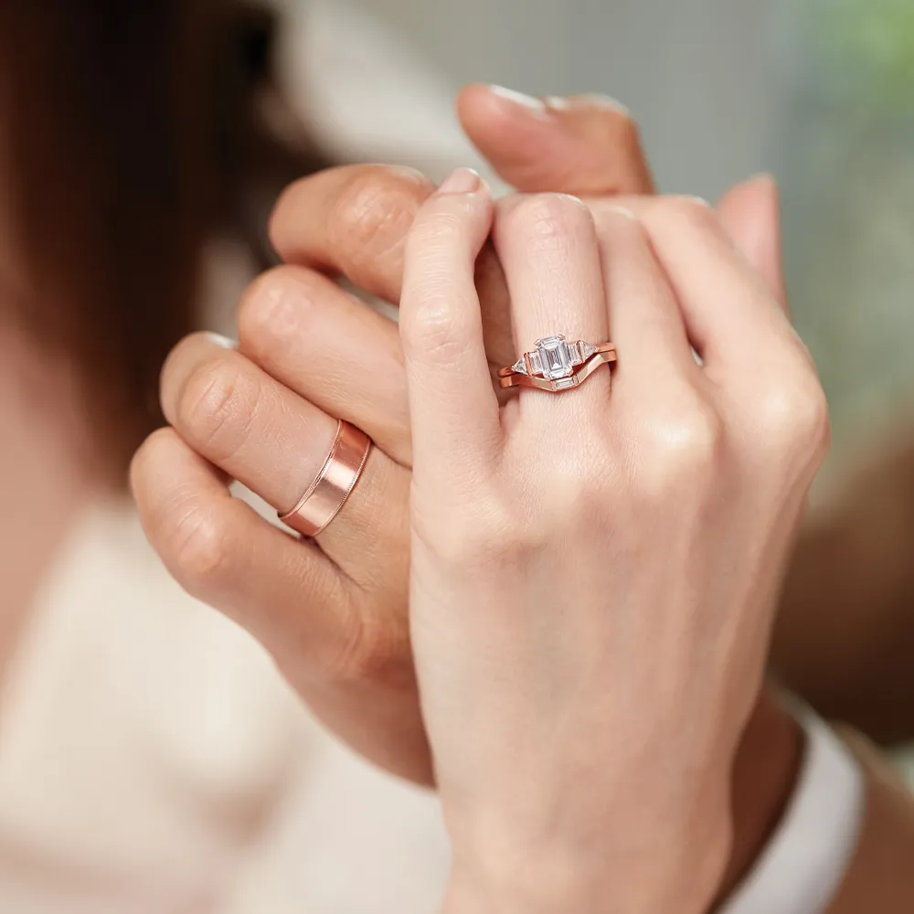 Two hands holding, one wearing a diamond engagement ring and the other a simple rose gold band.