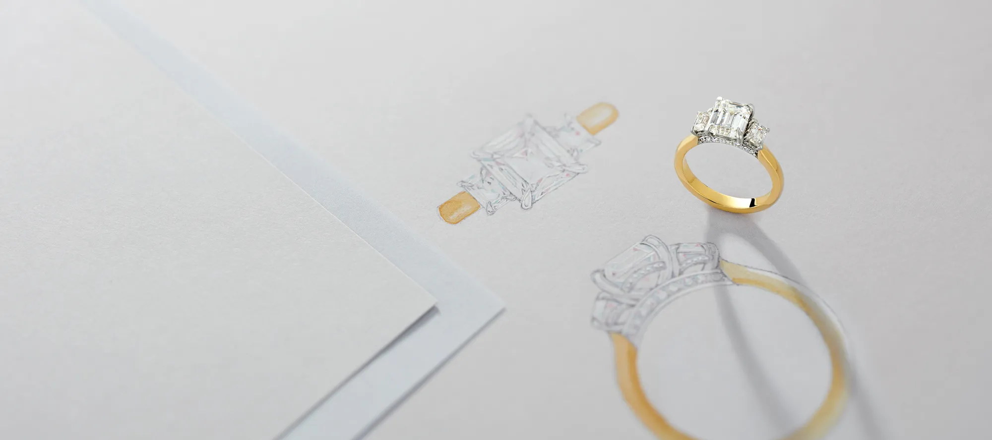 An elegant engagement ring beside its sketch on paper, showcasing a diamond centerpiece and gold band.