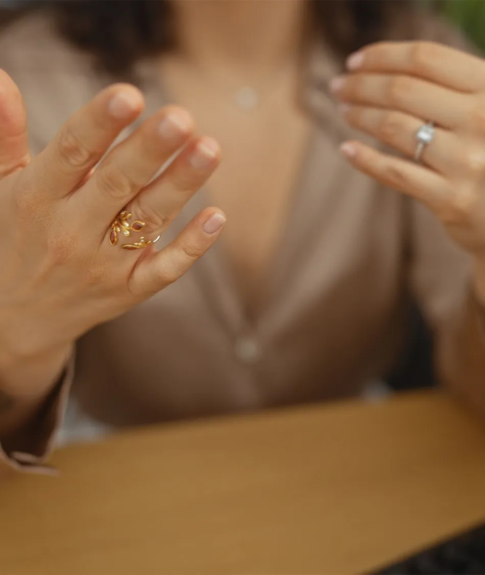 A woman shows her hand with a gold ring, wearing a beige blouse, with a diamond ring on her other hand.
