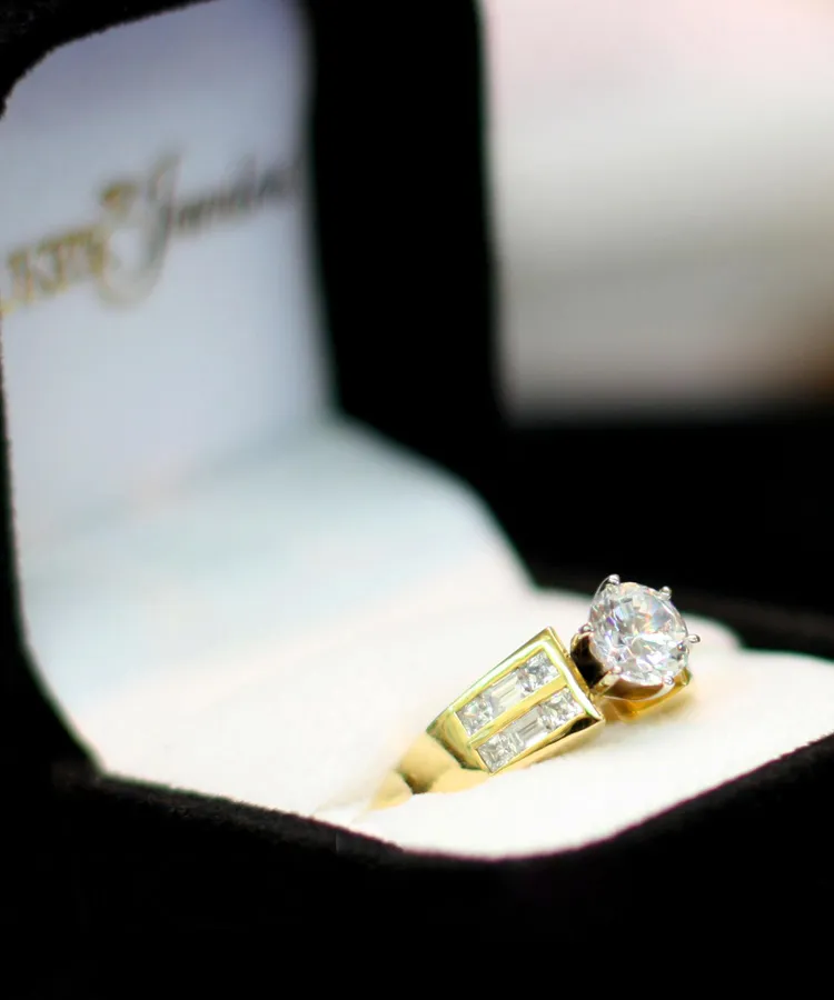 Gold engagement ring with a large diamond and smaller stones, displayed in a black velvet box.