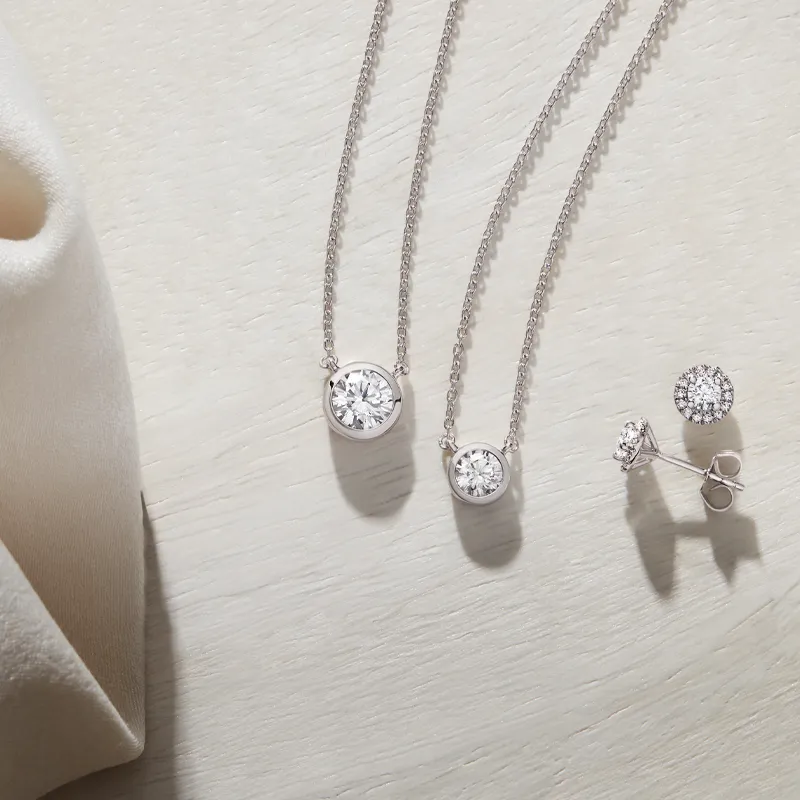 Two diamond necklaces and a pair of diamond stud earrings on a light wooden surface.