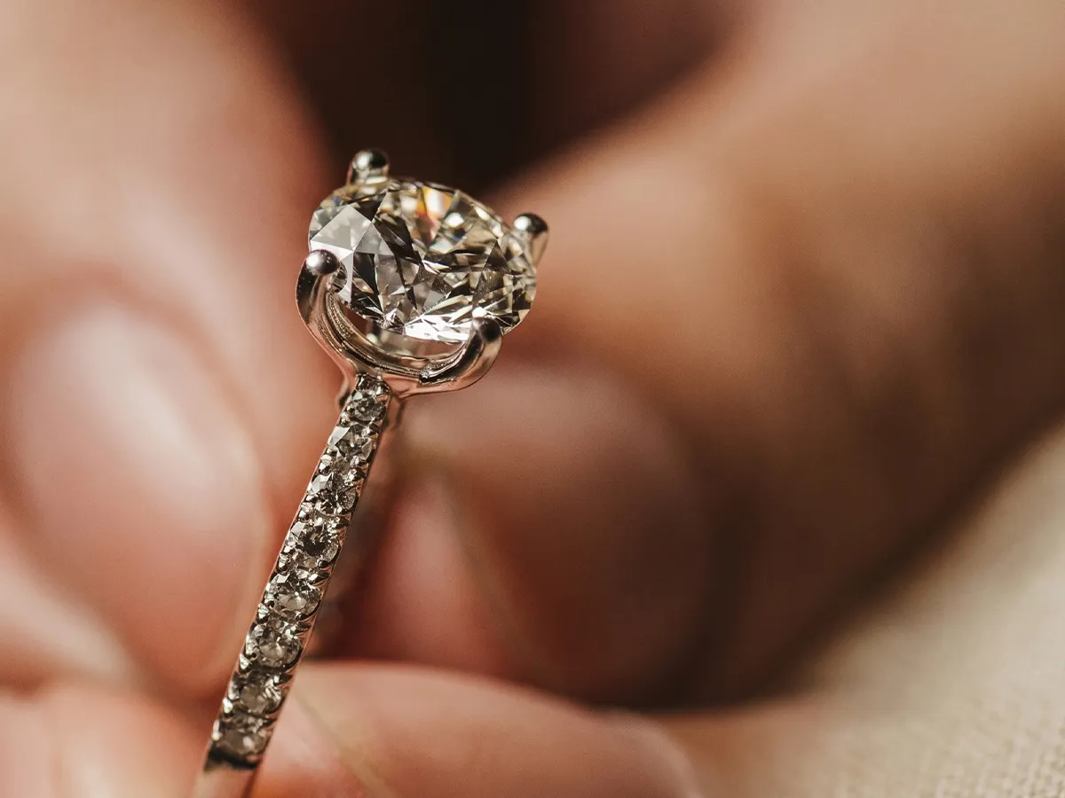 Close-up of a diamond engagement ring held between fingers. Book a Consultation.