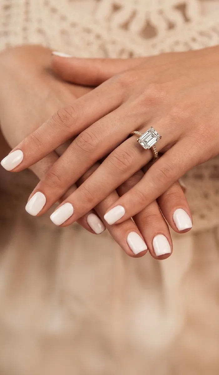 Hands with white nails displaying an emerald-cut diamond ring. Book a Virtual Consultation.