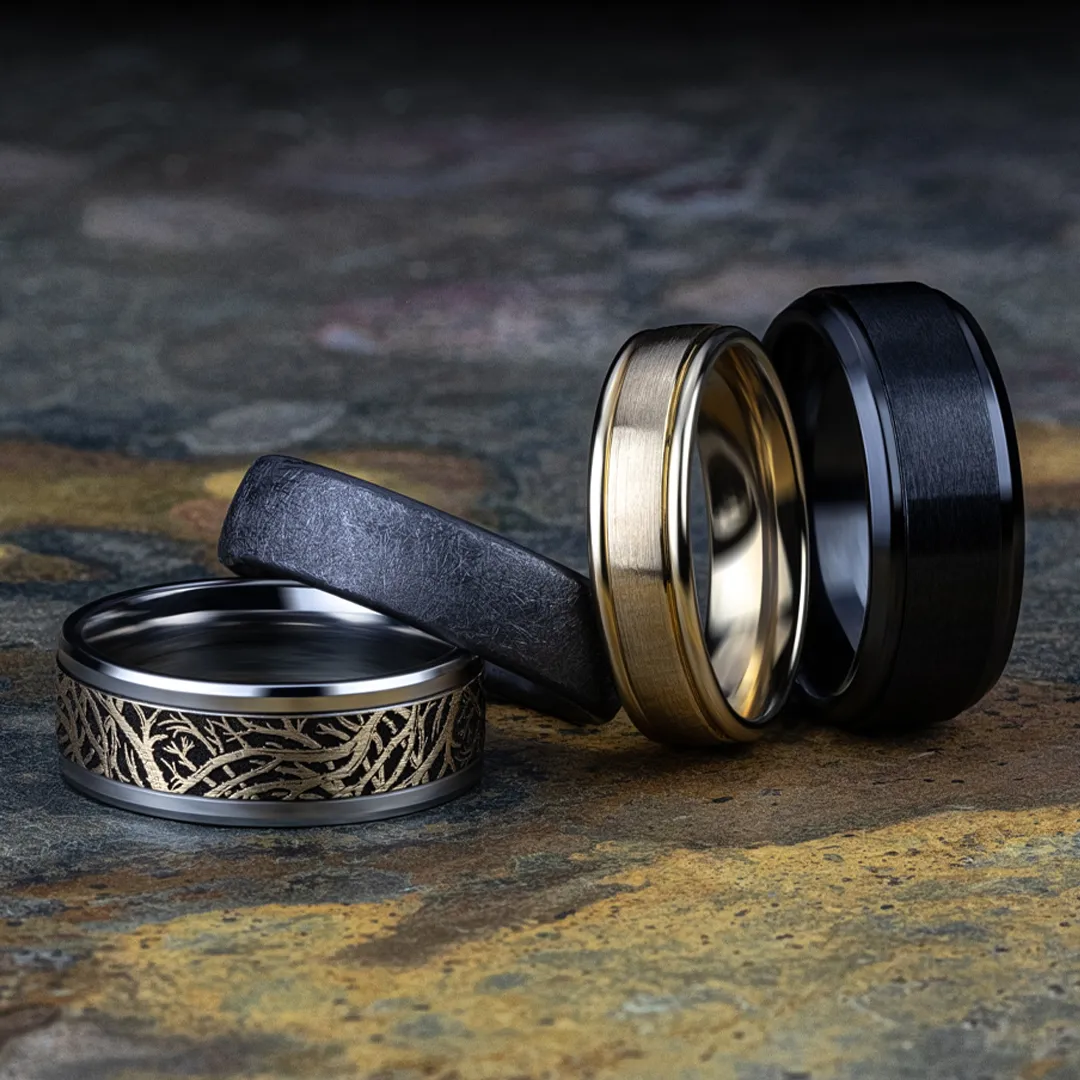 Four stylish rings in black, gold, and silver men's band designs displayed on a textured surface. Shop at Falls Jewelers.
