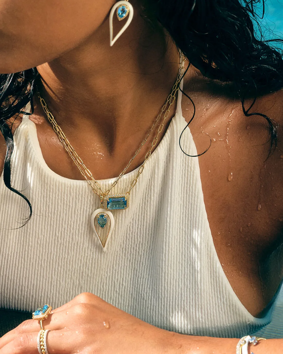 Woman wearing layered gold necklaces and earrings with blue gemstones, in a white top.