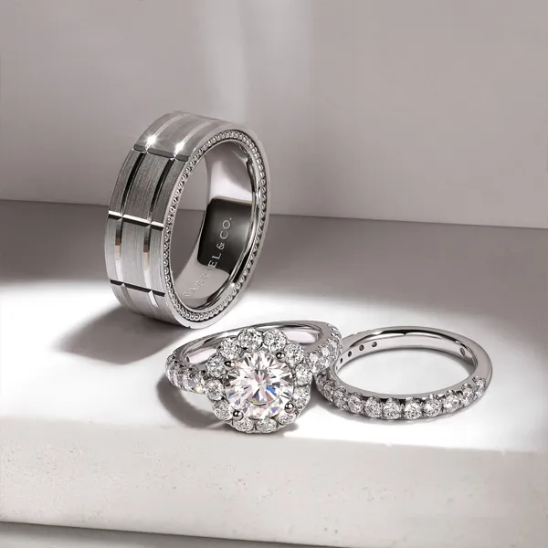 Three elegant wedding rings displayed on a surface, featuring a diamond halo, a simple band, and a textured band. Shop Wedding B