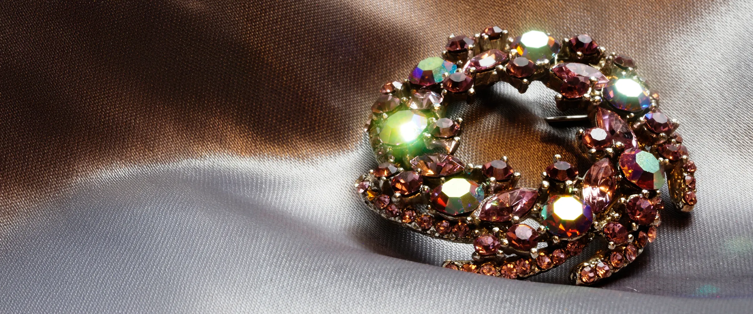 Sparkling brooch with pink and green gemstones on a silky fabric background.