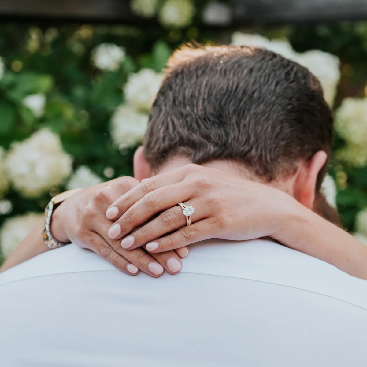 Engaged couple embracing, showcasing a diamond ring against a backdrop of white flowers.