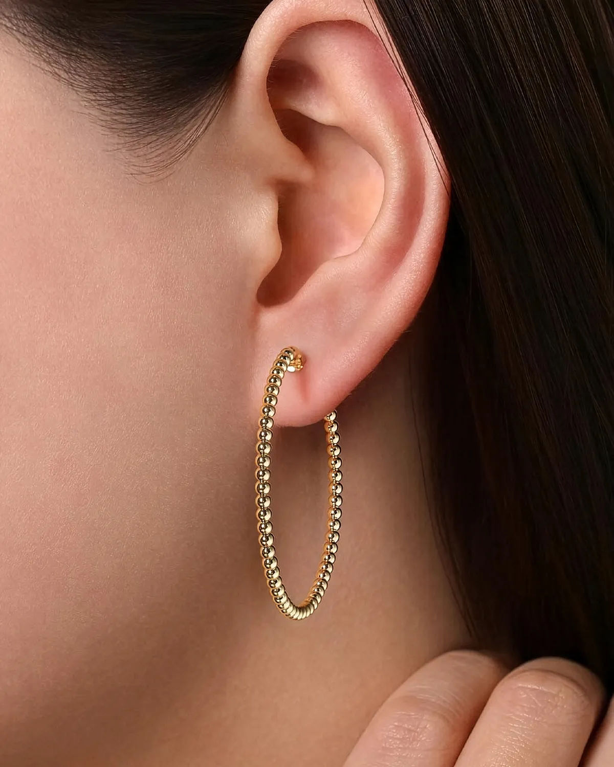 Gold hoop earrings with a beaded design, worn on a model's ear.