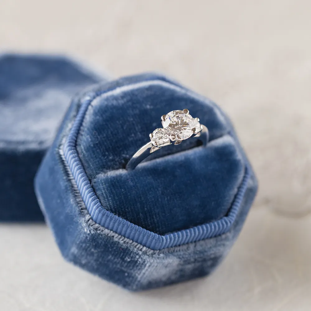 Elegant engagement ring with a central diamond and side stones, displayed in a blue velvet hexagonal box.