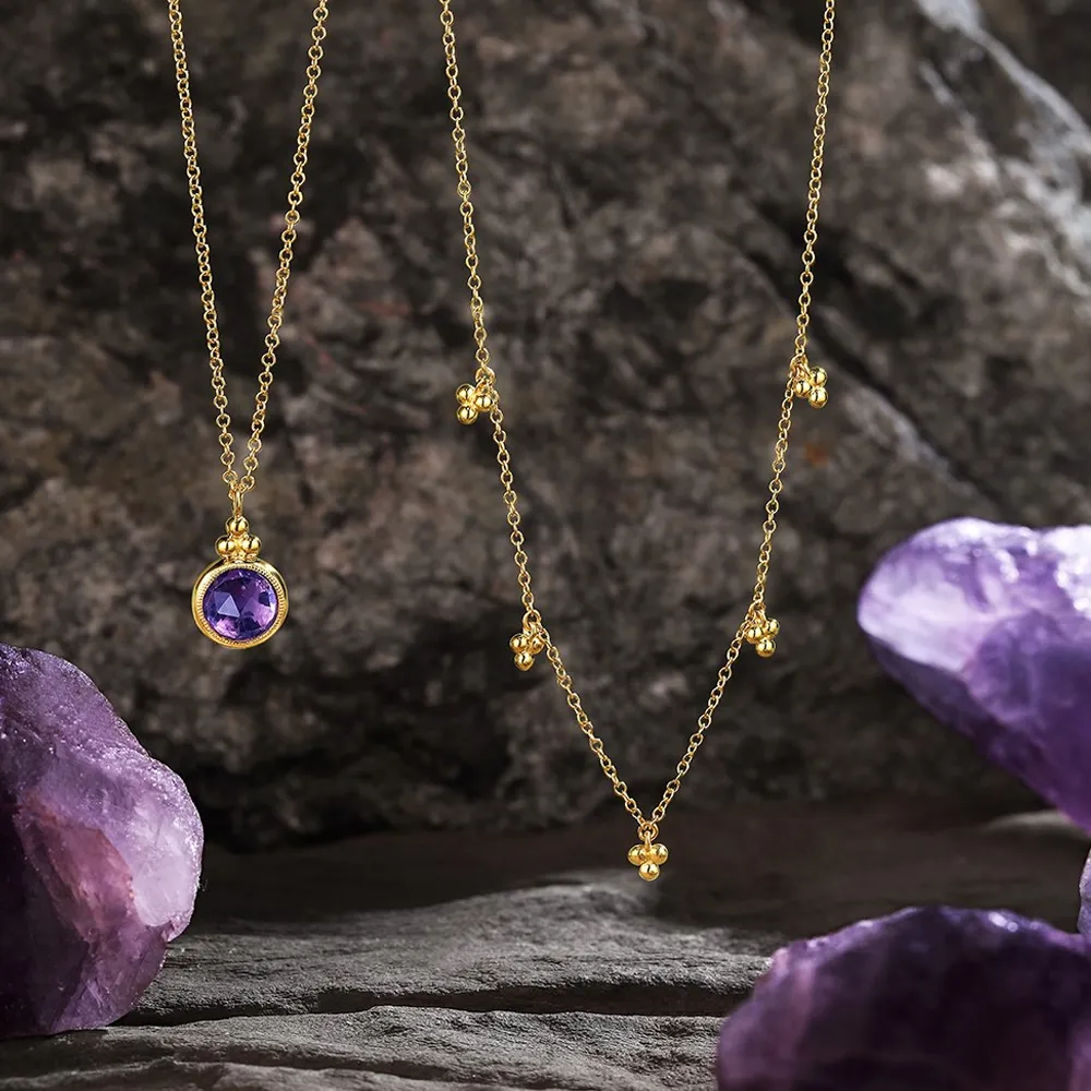 Gold necklaces with amethyst stones displayed on a rocky surface, surrounded by purple amethyst crystals.