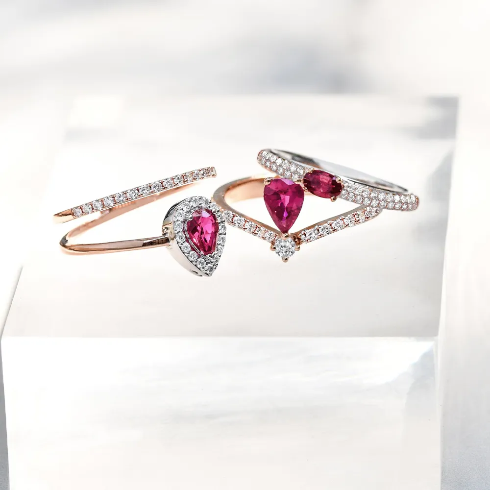 Three elegant ruby rings with diamonds, showcasing heart and pear shapes, displayed on a clear surface.