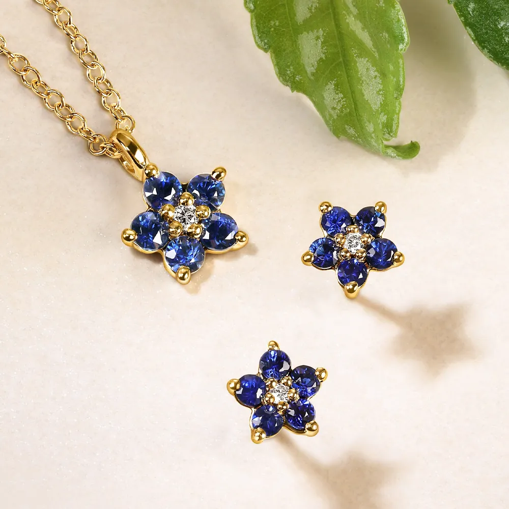 Gold flower pendant and earrings set with blue sapphires and diamonds, displayed on a light surface with green leaves.