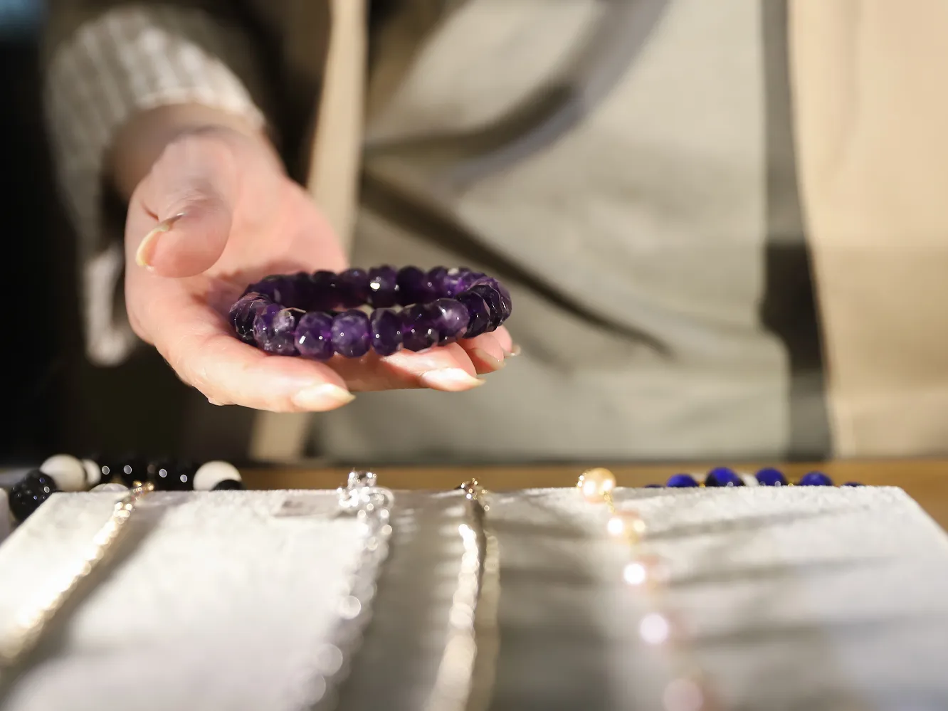 A hand holding a purple amethyst bracelet over a display of various jewelry pieces. Visit Flora Gems for more.