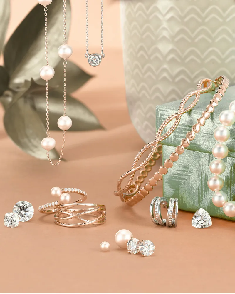 Jewellery displayed on light blue gift box, gold rings, loose diamonds and pearls scattered around with gold necklaces hanging. 