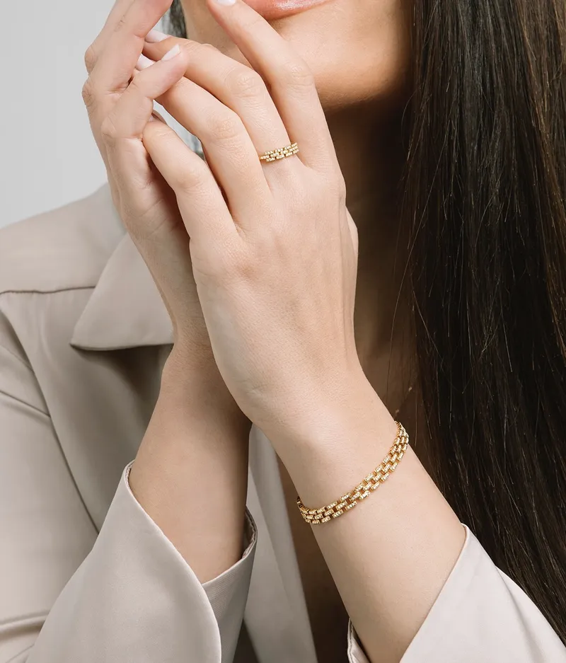 Model showcasing gold jewelry, including a ring and bracelet wearing a tan jacket.