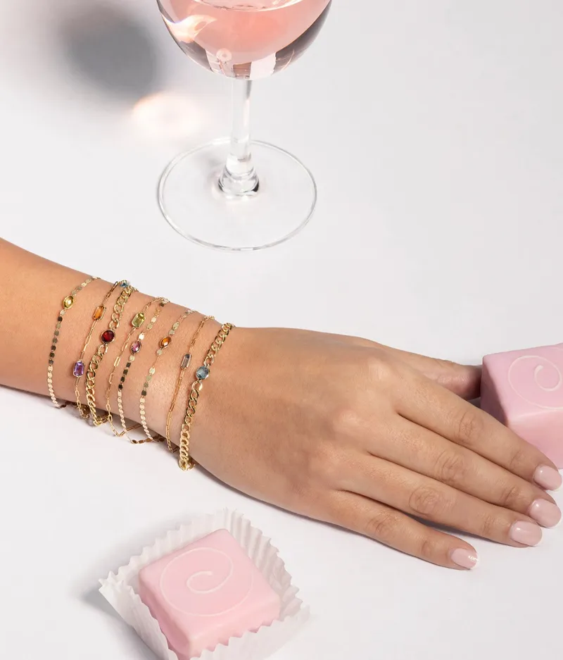 Elegant hand adorned with colorful gold bracelets, pink drink, and sweets. Click to Shop Now at Futer Bros Jewelers.